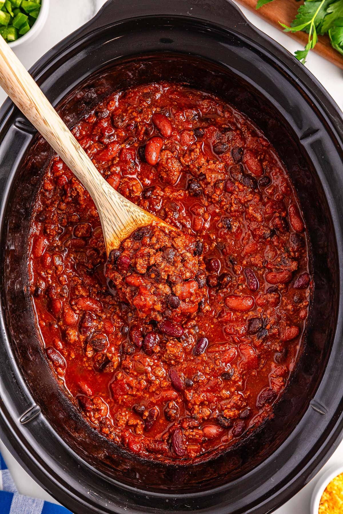 A slow cooker filled with hearty slow cooker chili, brimming with beans and ground meat, as a wooden spoon scoops out a steaming, savory helping.