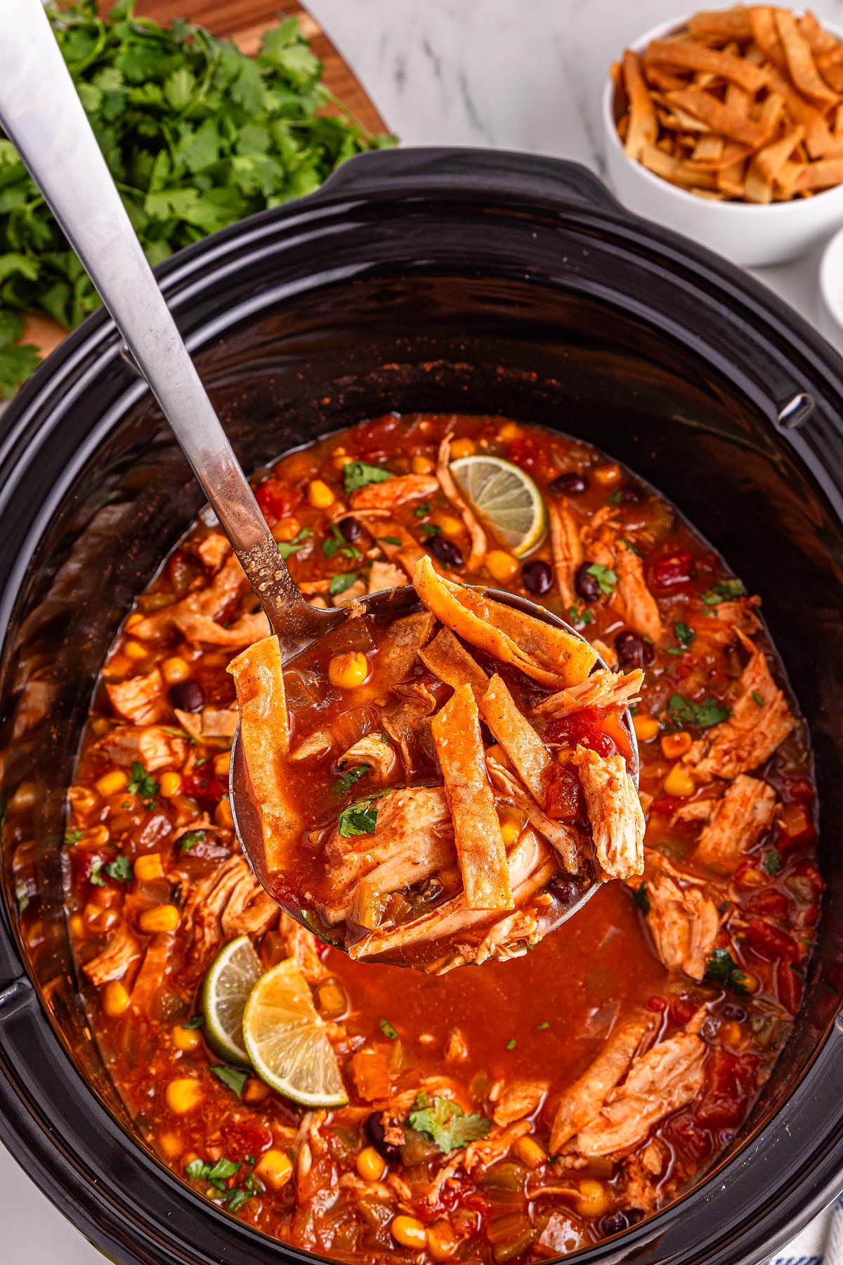 Ladle lifting Slow Cooker Chicken Tortilla Soup with vegetables and lime from a slow cooker, garnished with fresh herbs.