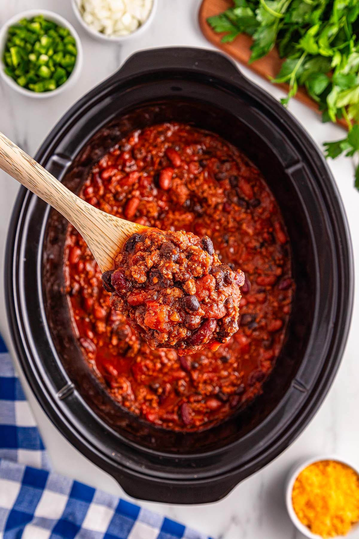 A wooden spoon lifts hearty slow cooker chili from a crockpot, surrounded by bowls of chopped veggies and cheese.