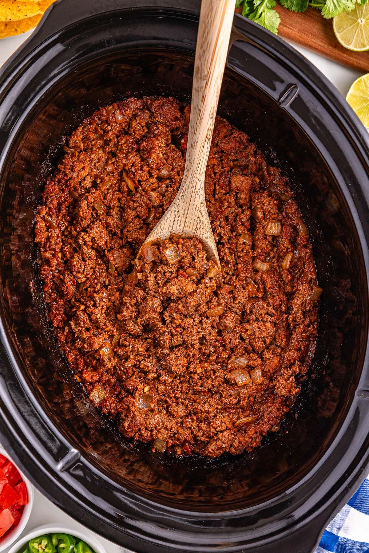 Slow Cooker Taco Meat is shown in a slow cooker with a wooden spoon, surrounded by bowls of fresh toppings.