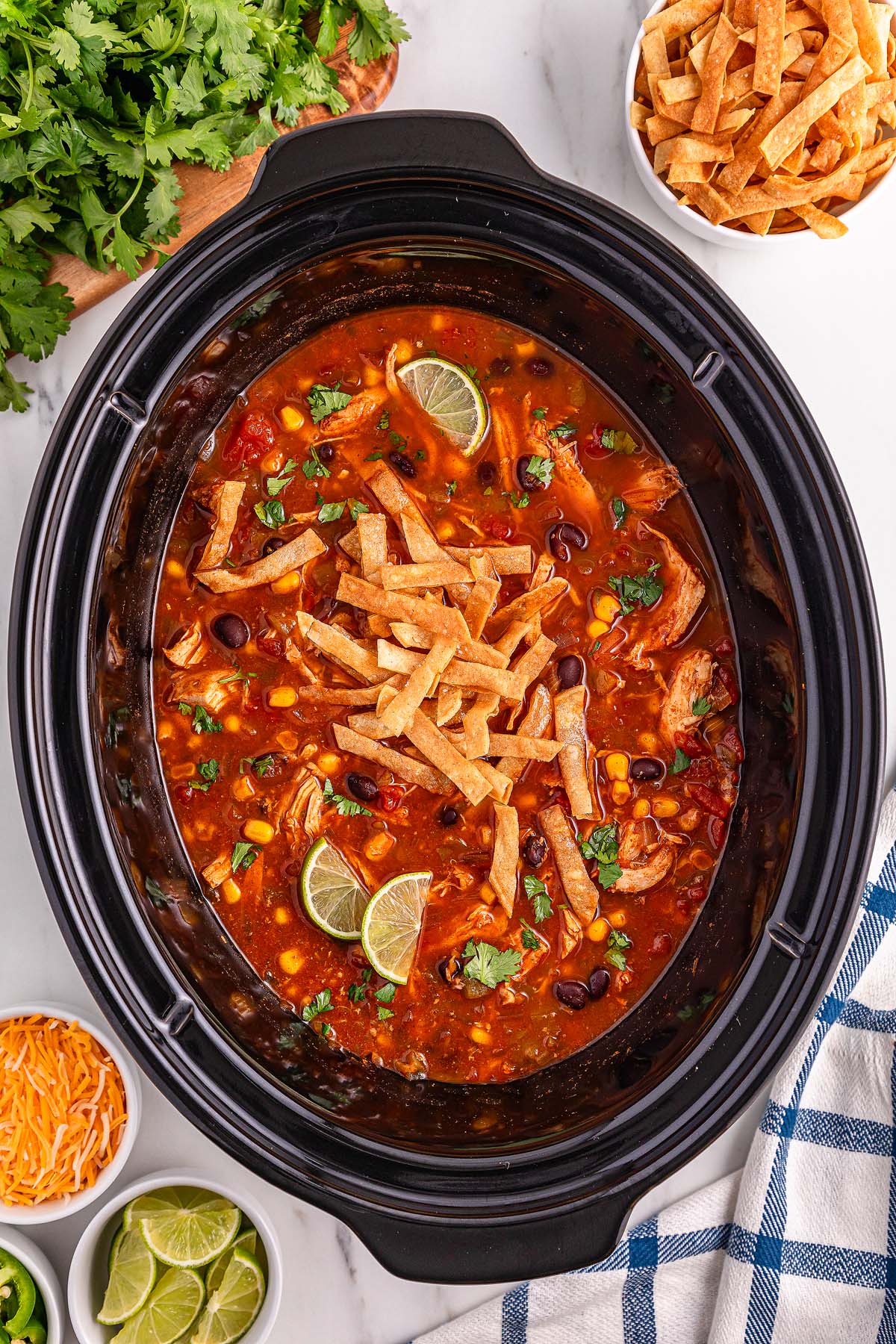 A slow cooker filled with flavorful Slow Cooker Chicken Tortilla Soup, topped with crispy tortilla strips and fresh lime, surrounded by an array of delicious toppings.