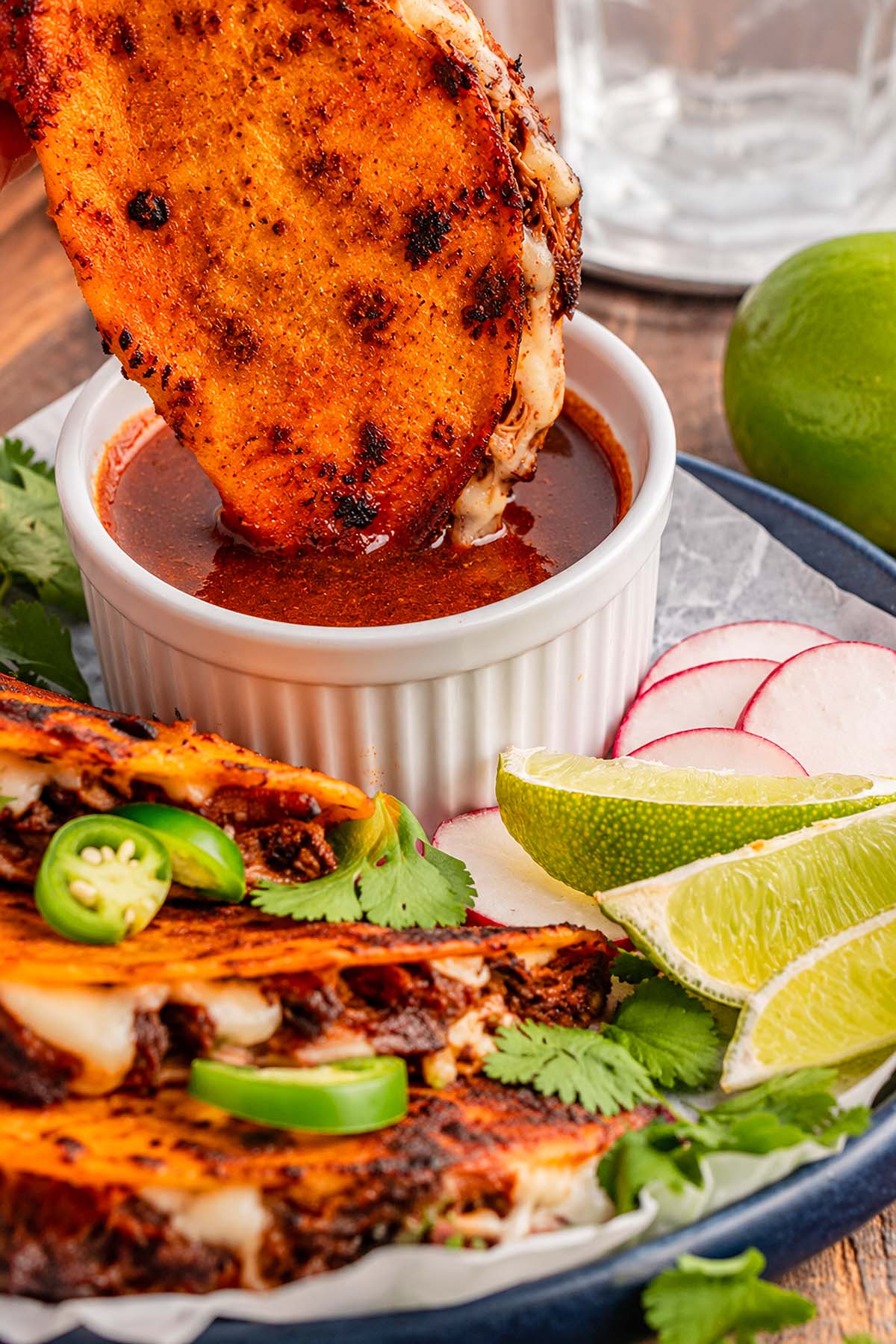 Grilled taco dipped in red sauce made with flavorful slow cooker birria, served with lime wedges, radish slices, jalapeño, and cilantro on the side.