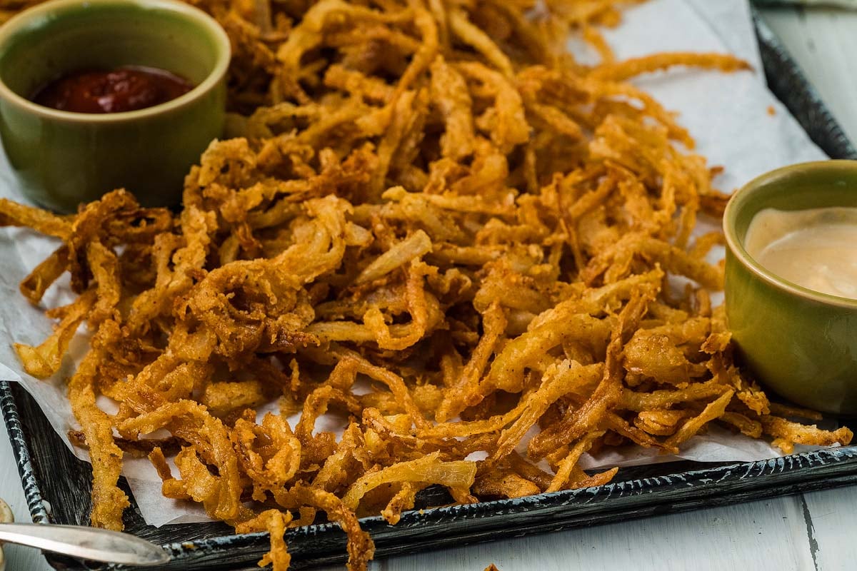 Crispy onion strings are served on parchment, accompanied by ketchup and a creamy dipping sauce in small bowls.