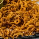 Golden crispy onion strings piled high on a tray, served with flavorful dipping sauces in small bowls on the side.