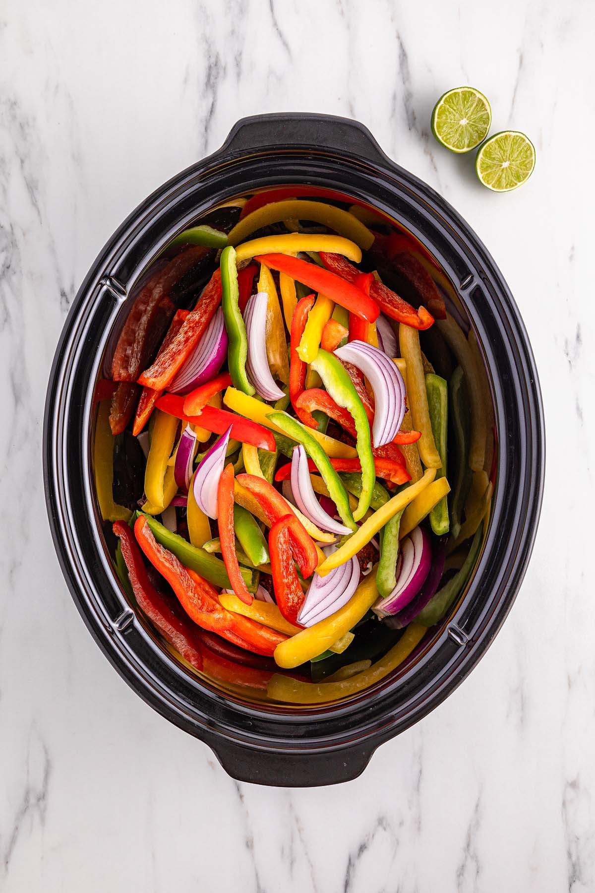 Sliced bell peppers and red onions in a slow cooker on a marble surface, ready to create flavorful Slow Cooker Chicken Fajitas, with two lime halves beside it.