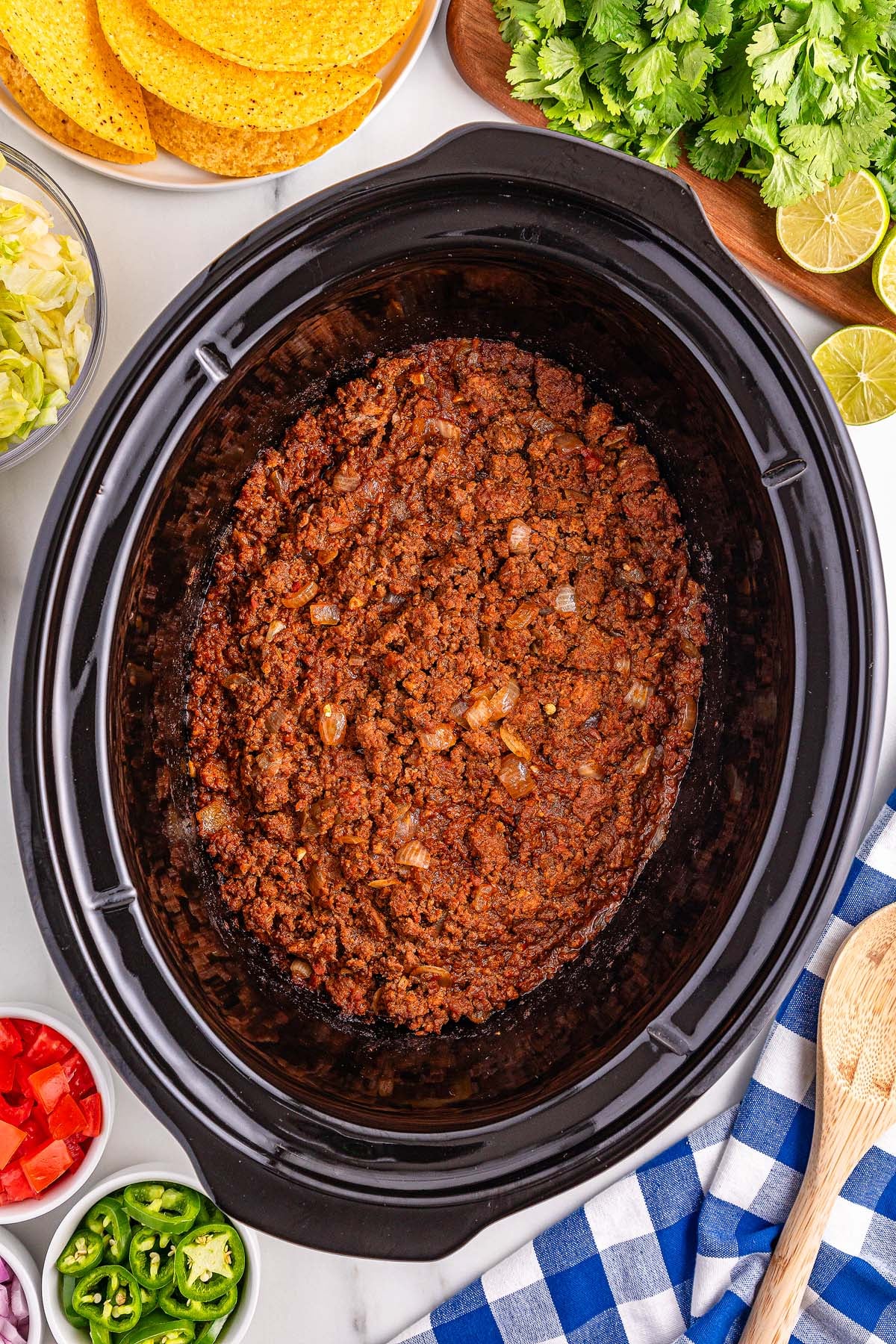 Slow Cooker Taco Meat simmers to perfection in your slow cooker, creating a flavorful ground beef taco filling surrounded by crispy taco shells, fresh veggies, cilantro, lime wedges, and a wooden spoon for serving.