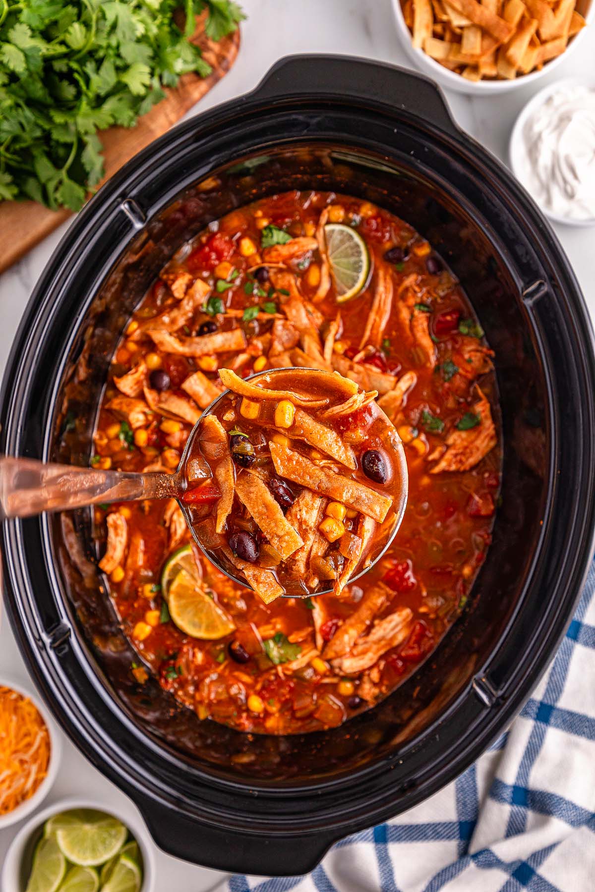 A ladle scoops up hearty Slow Cooker Chicken Tortilla Soup, brimming with beans, corn, and fresh lime from a simmering slow cooker.
