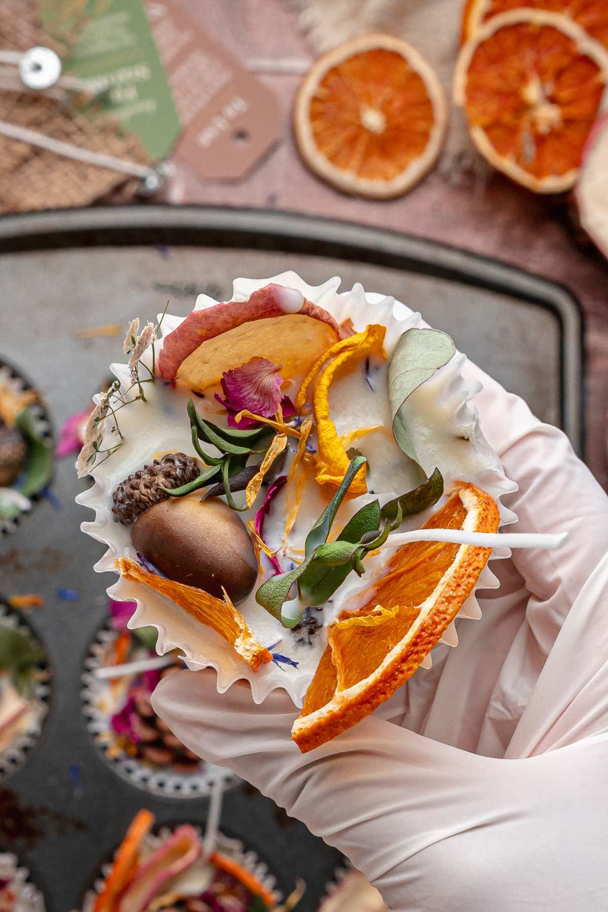 A gloved hand holds a fire starter topped with dried orange slices, flowers, leaves, and botanicals.