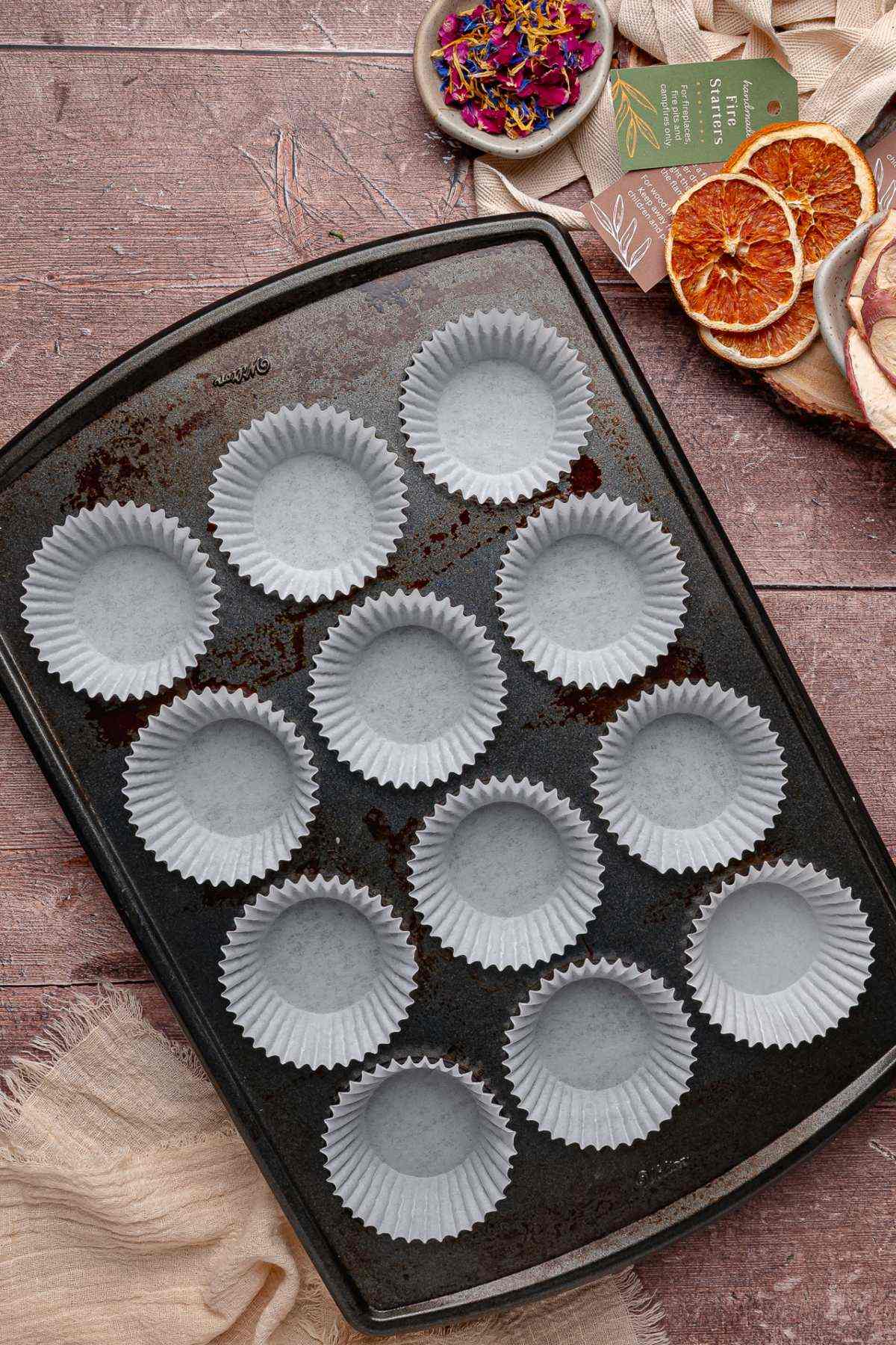 A muffin tin with twelve white paper liners on a wooden table, surrounded by dried fruit and flowers.
