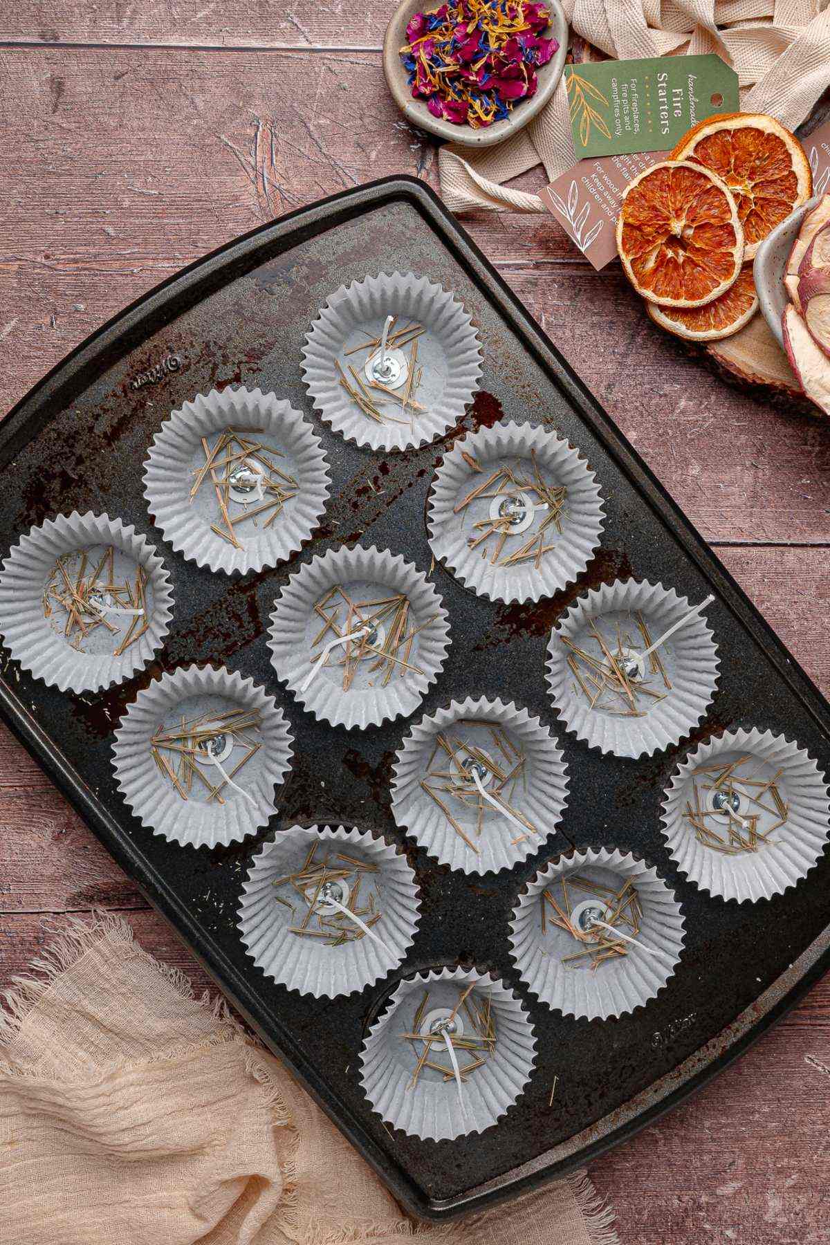 A baking tray with paper liners filled with dried herbs, with dried oranges, wicks and flowers nearby.