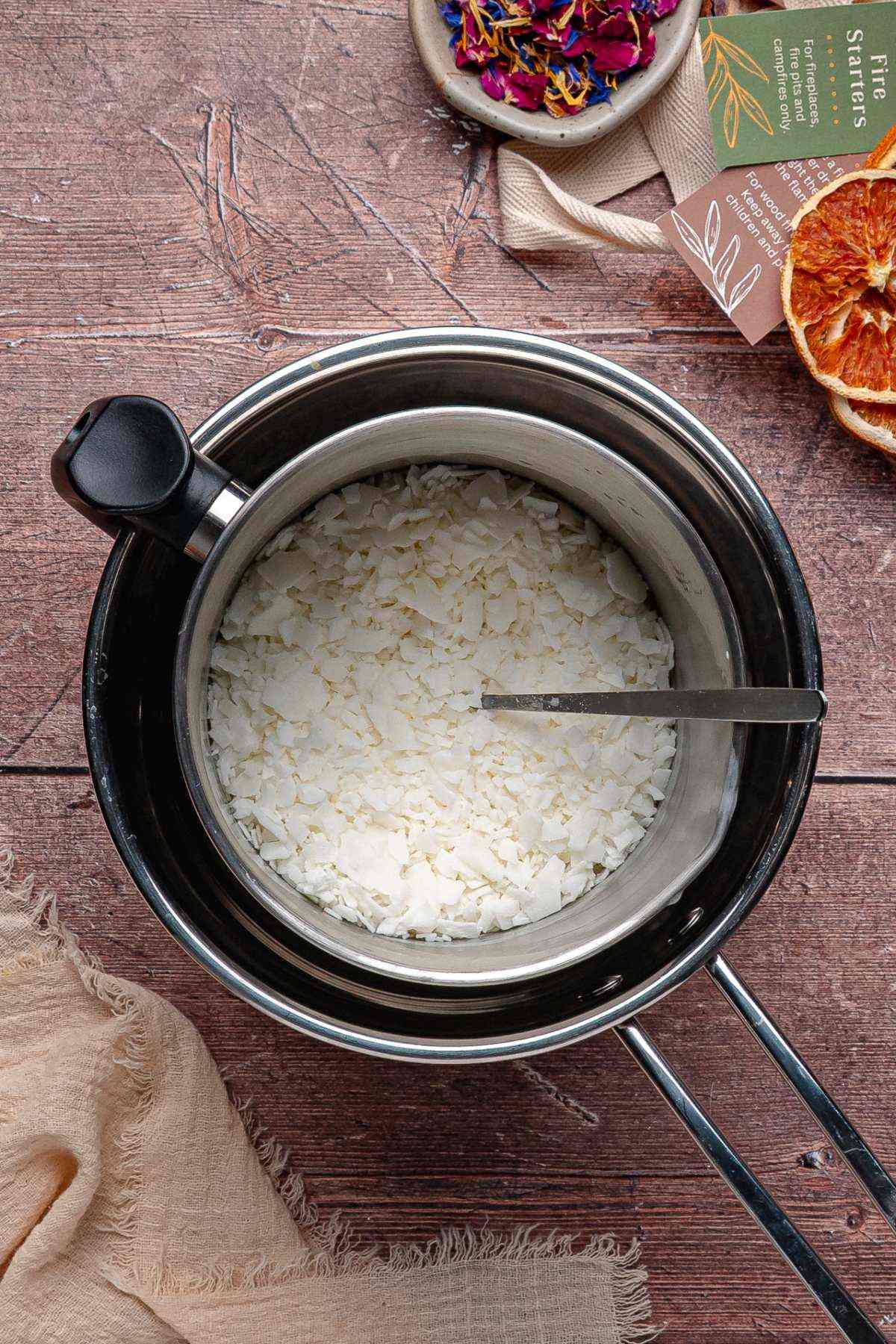 Metal pot with white wax flakes melting inside a double boiler, with a spoon resting in the pot.