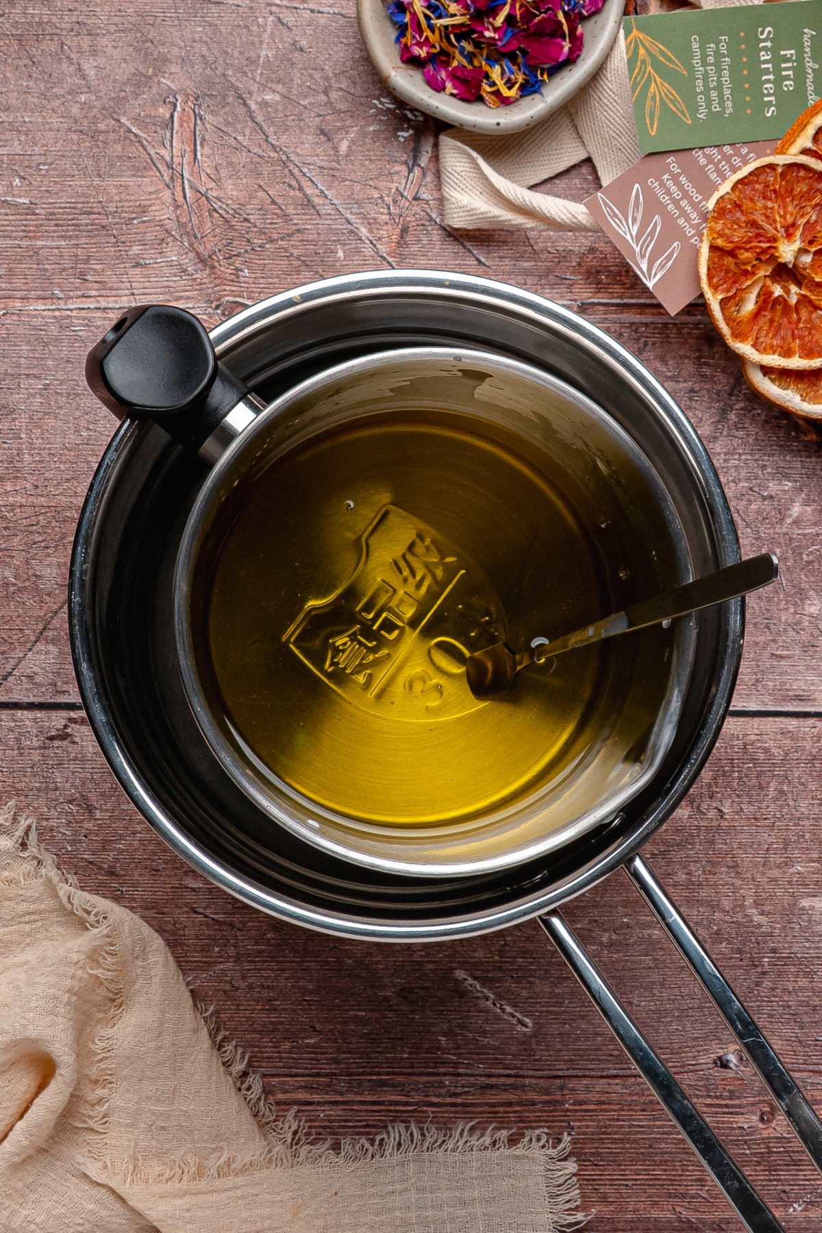 A metal pot with melted wax and a thermometer inside, set on a wooden surface with dried oranges nearby.