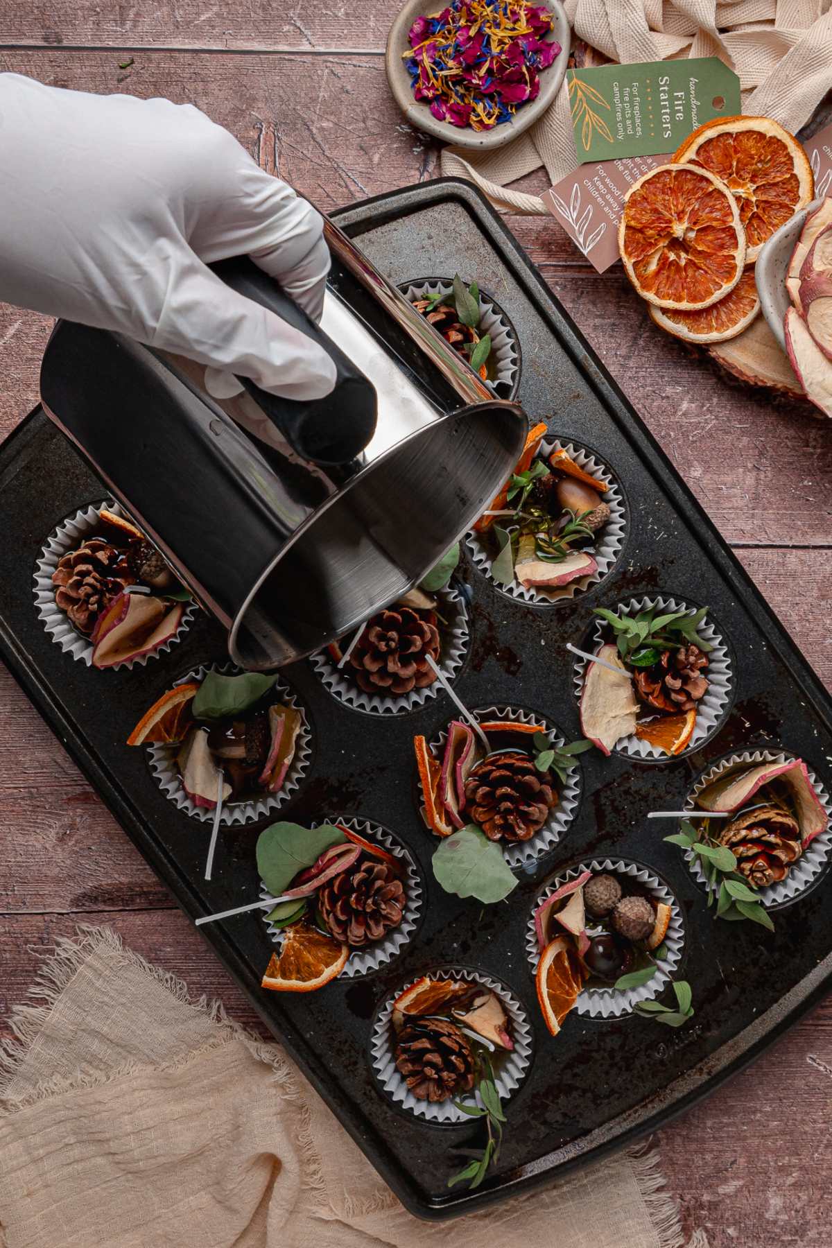 A gloved hand pours liquid wax into muffin tins filled with pinecones, leaves, and dried fruits on a baking tray.