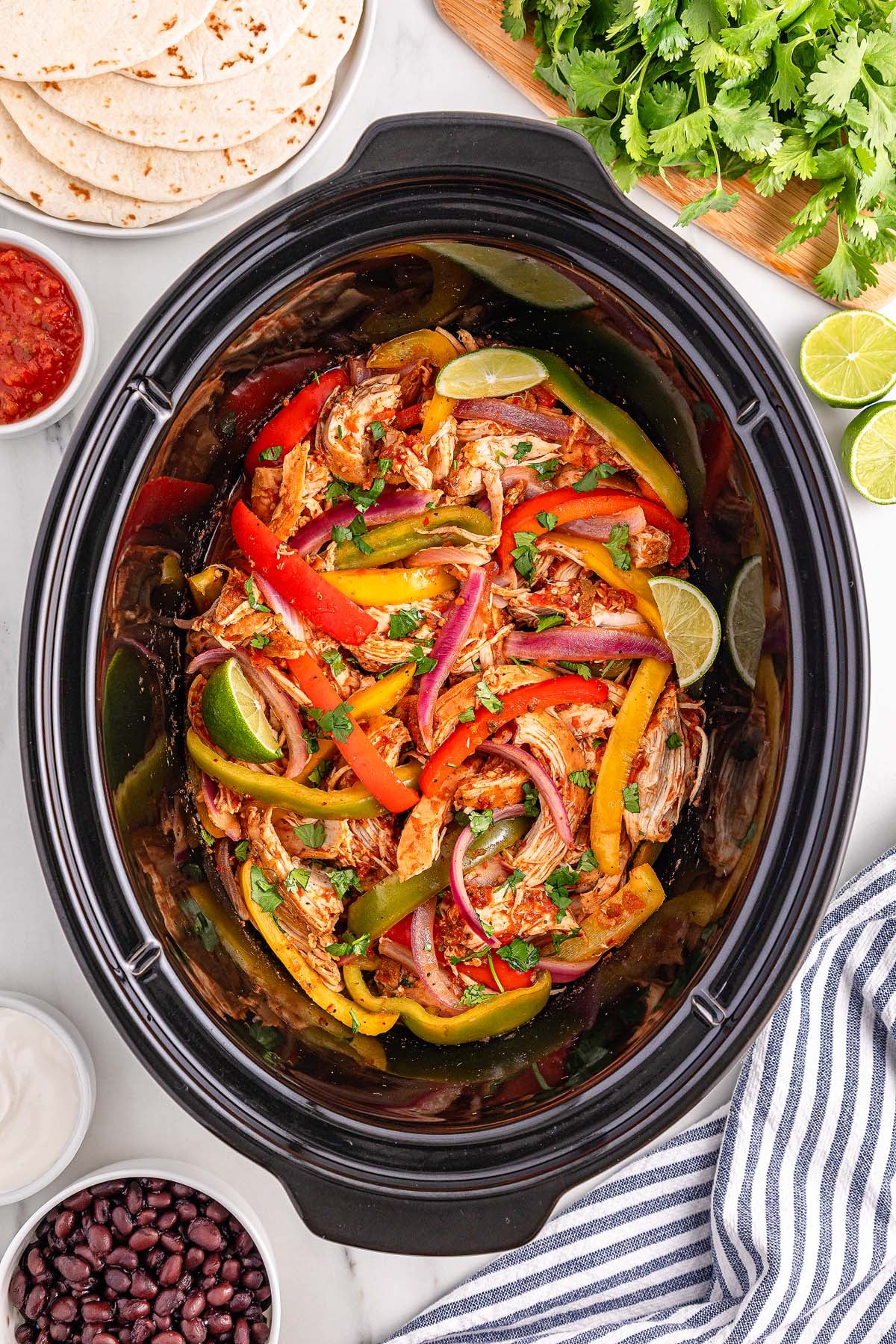 Slow Cooker Chicken Fajitas simmer in a slow cooker, surrounded by warm tortillas, fresh limes, cilantro, and small bowls of flavorful sides.