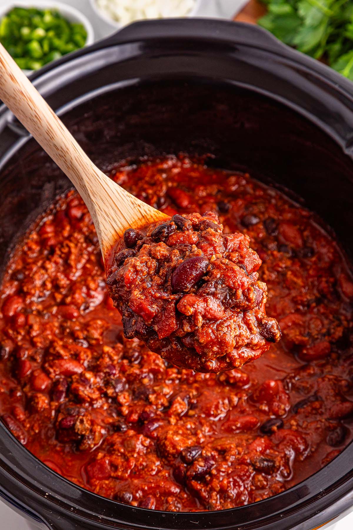 A wooden spoon lifts chunky slow cooker chili with beans and ground beef from a black slow cooker.