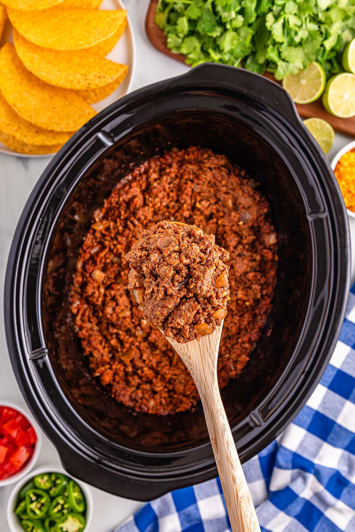 A wooden spoon holds flavorful Slow Cooker Taco Meat over a slow cooker, surrounded by crispy taco shells and fresh toppings.