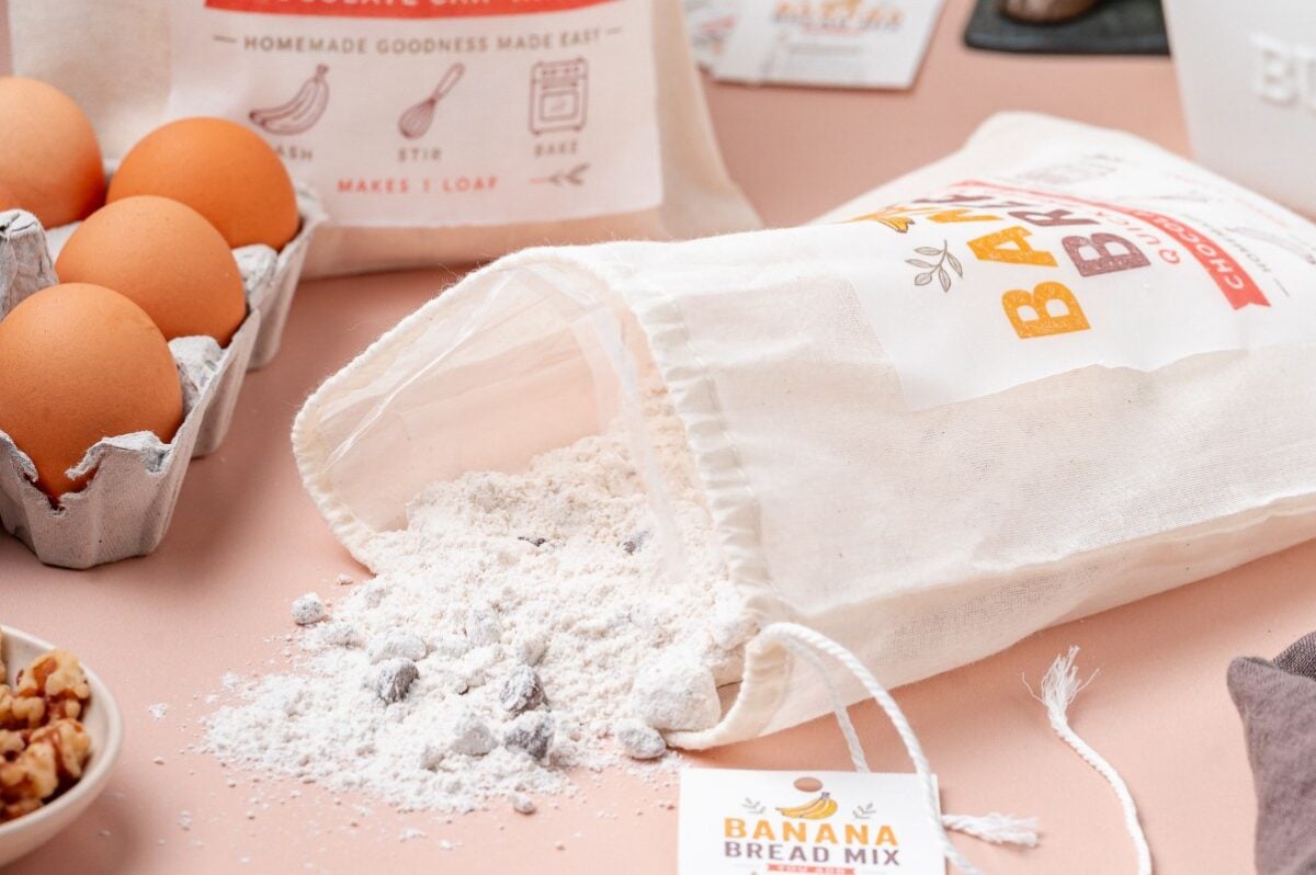 Homemade banana bread mix spilling from a cloth bag onto a counter with eggs, walnuts, and baking supplies nearby.