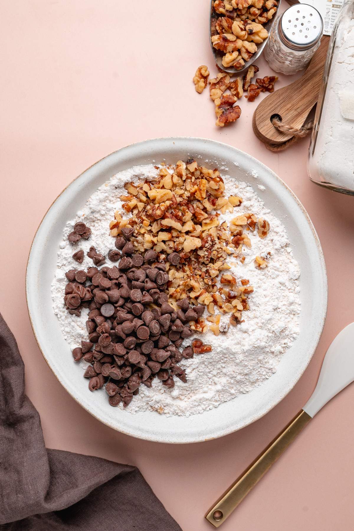 A bowl of flour with chocolate chips and chopped walnuts on top, surrounded by baking ingredients and utensils, is ready to become a delicious homemade banana bread mix.