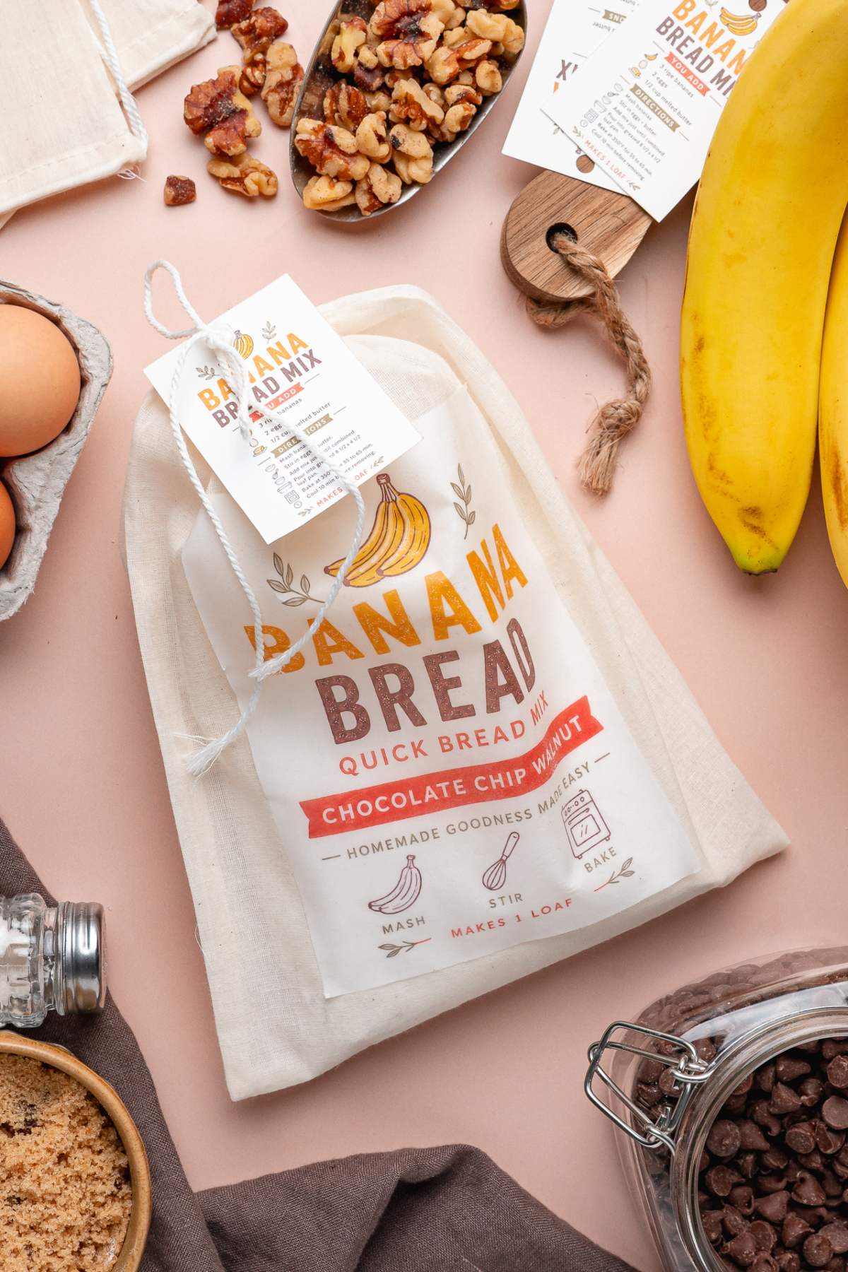 A homemade banana bread mix bag is surrounded by ingredients like bananas, eggs, walnuts, and chocolate chips on a table.
