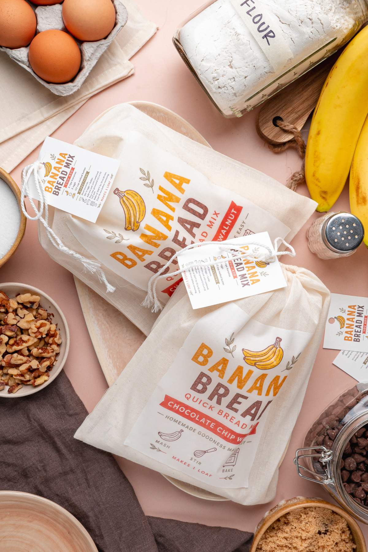 Homemade banana bread mix bags surrounded by baking ingredients like eggs, bananas, flour, nuts, and chocolate chips.