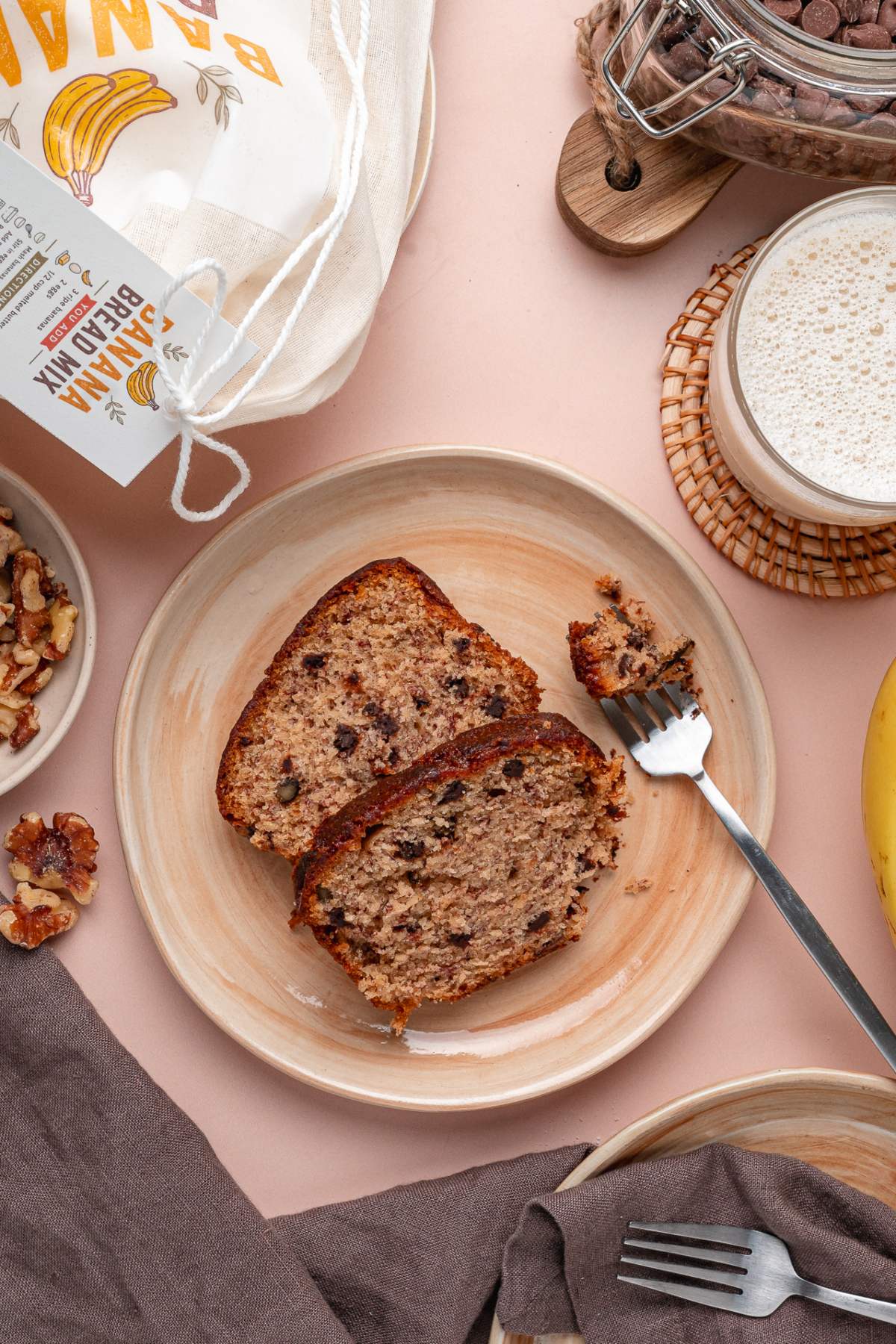 Two slices of banana bread with chocolate chips made from a homemade banana bread mix sit on a plate, surrounded by a fork, walnuts, and a glass of milk nearby.