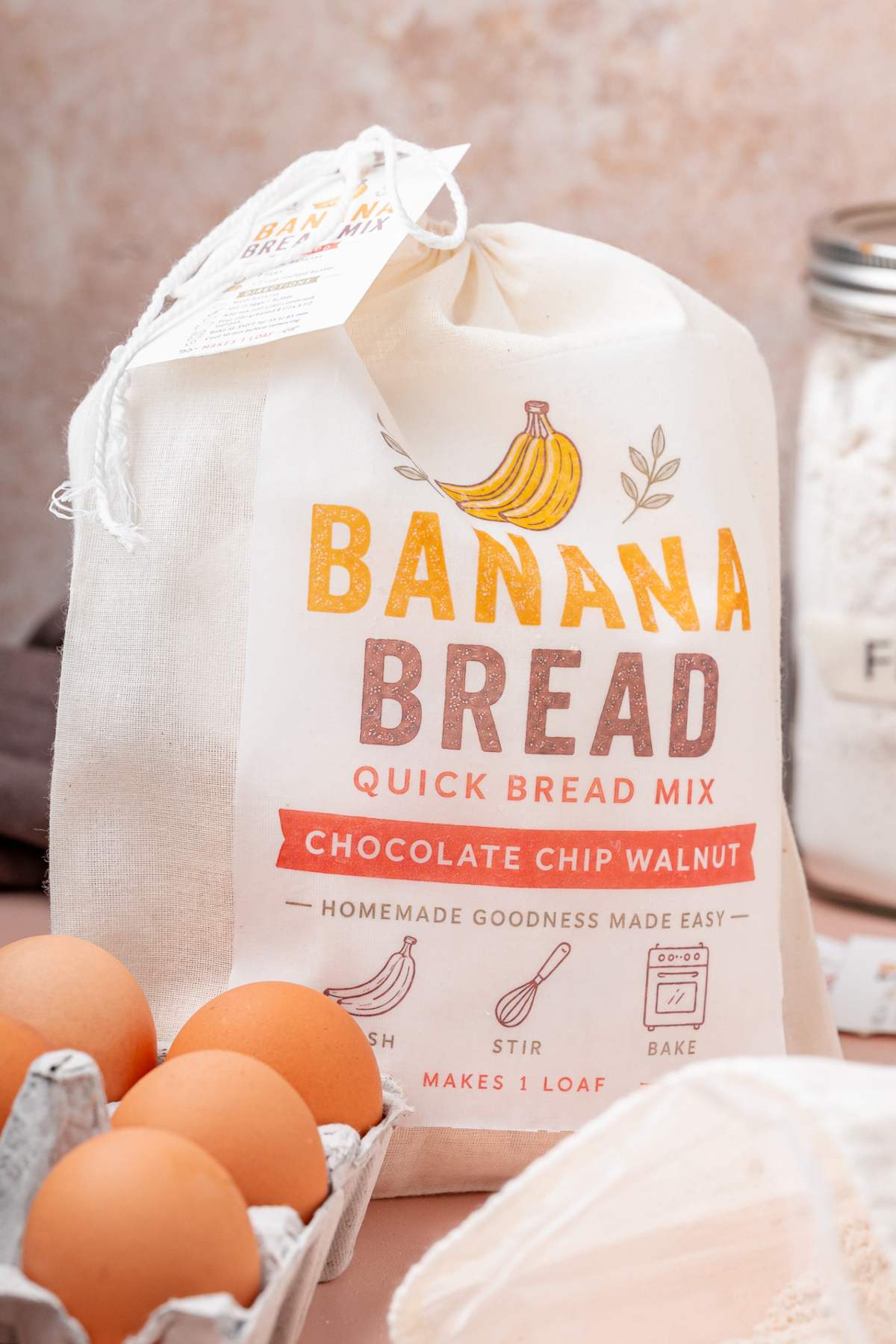 A bag labeled "Homemade Banana Bread Mix" sits on a kitchen counter, with eggs and a flour jar nearby, ready for baking.