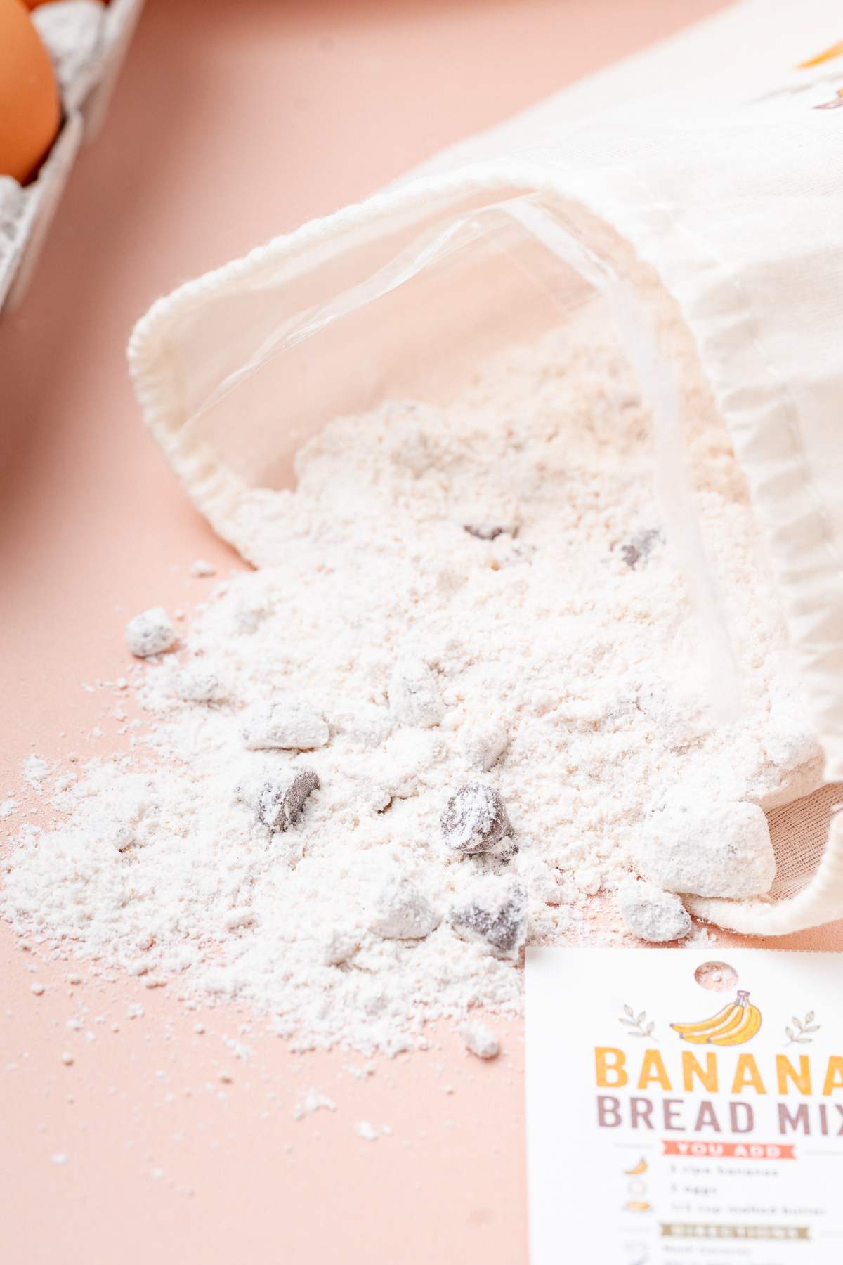 Homemade banana bread mix with chocolate chips spills from a fabric bag onto a light pink surface.