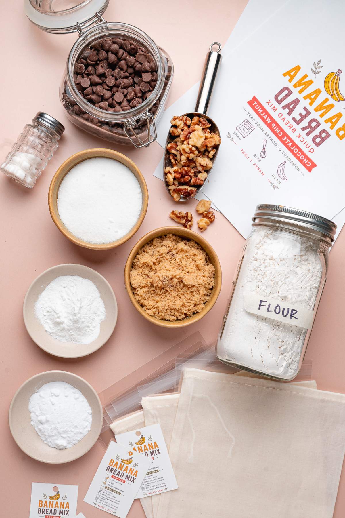 Baking ingredients including flour, sugars, chocolate chips, walnuts, homemade banana bread mix, and labeled recipe cards on a counter.