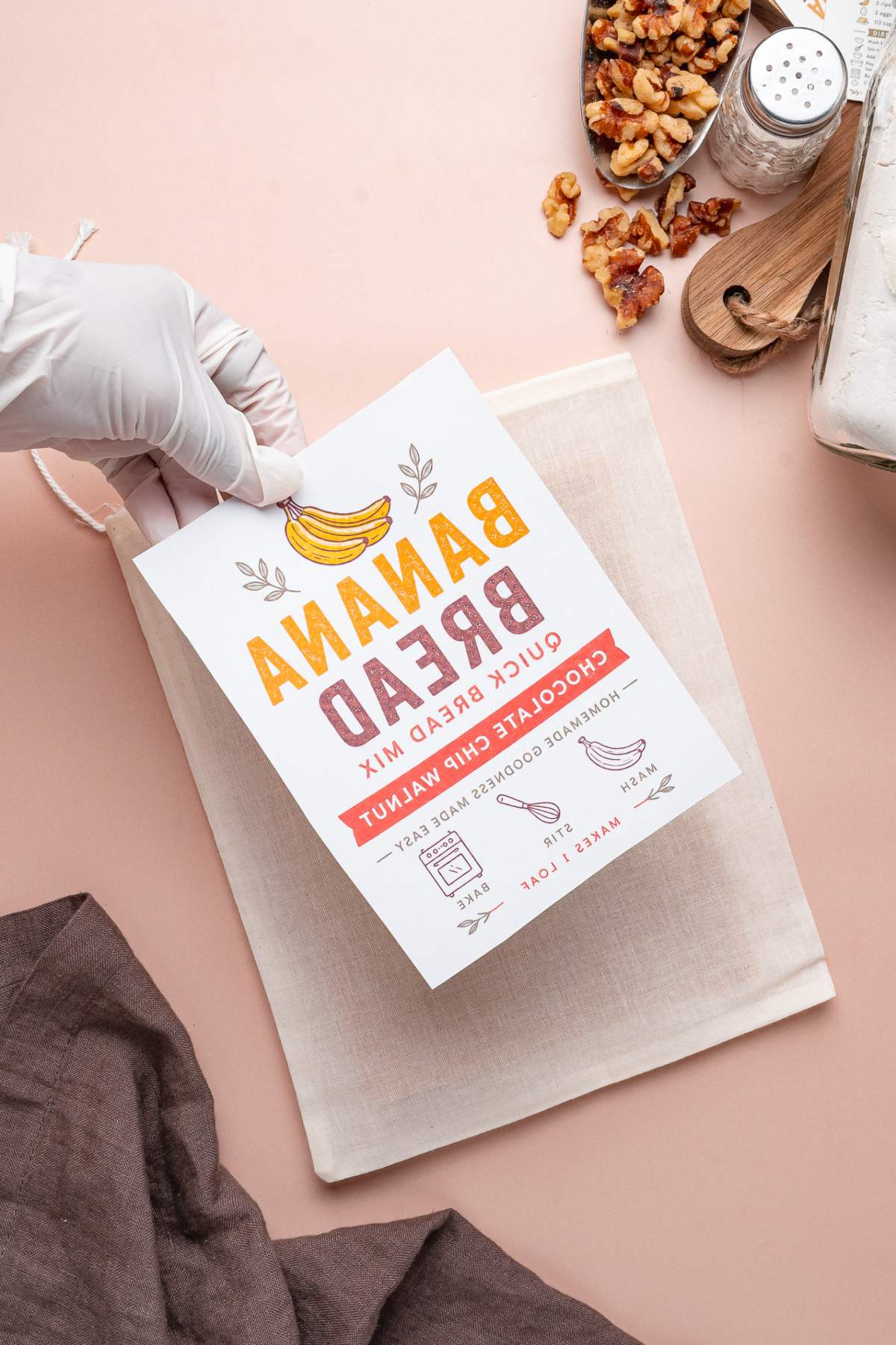 A gloved hand holds a banana bread recipe card above a cloth bag filled with homemade banana bread mix on a pink surface, with ingredients nearby.