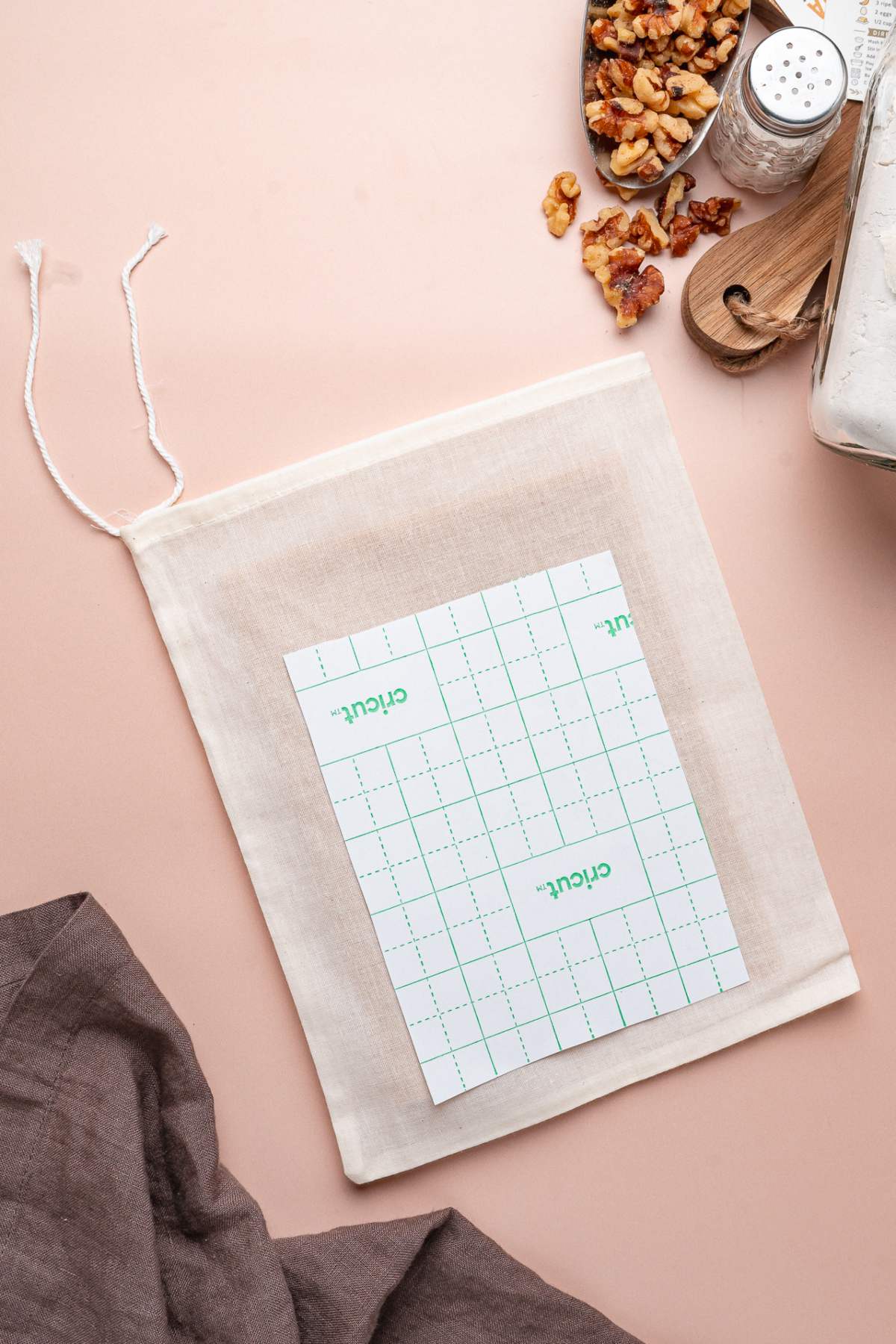 A notepad on a drawstring cloth bag, near walnuts, a gray cloth, homemade banana bread mix, and other baking ingredients on a pink surface.