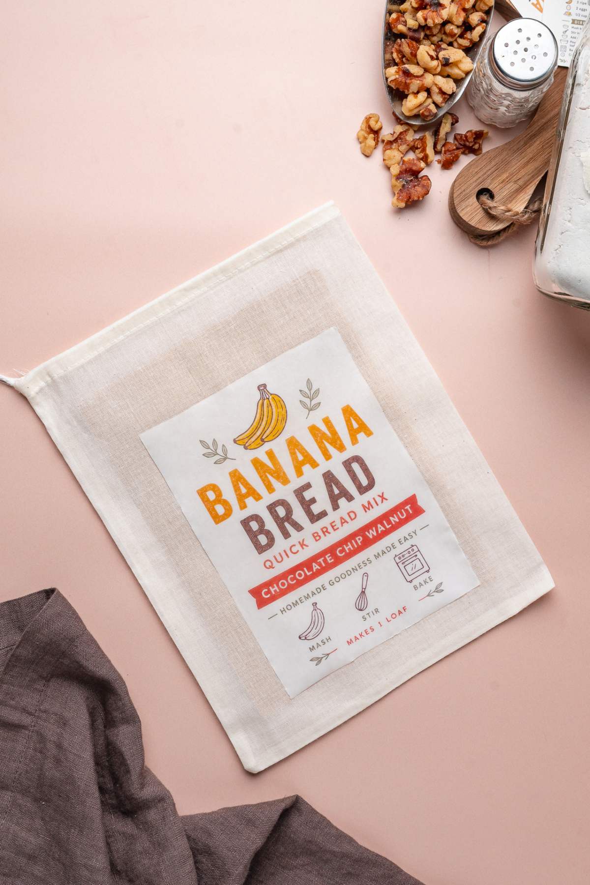 A homemade banana bread mix pouch labeled "Banana Bread Quick Bread Mix, Chocolate Chip Walnut" rests on a pink surface.