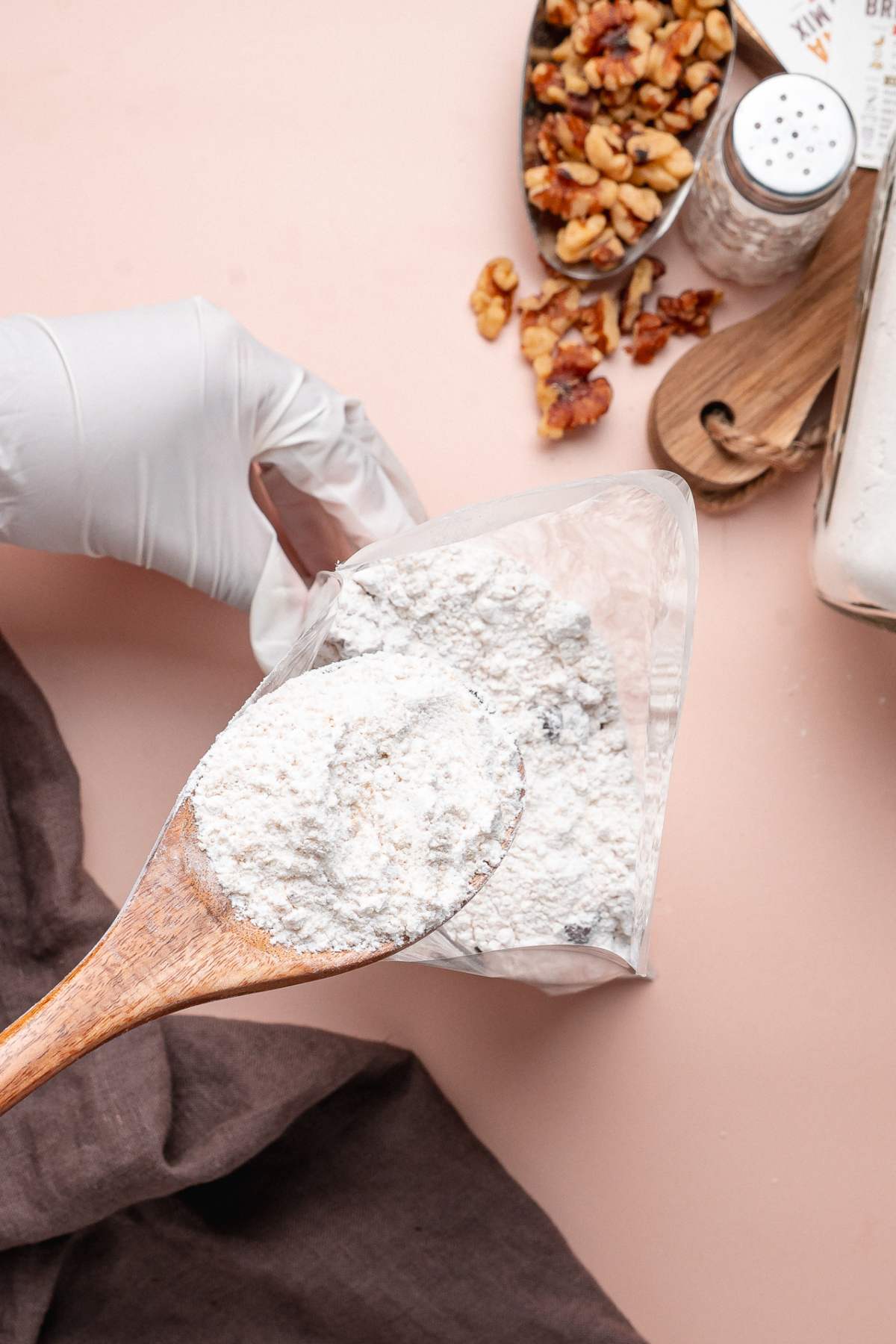 A gloved hand holds a bag of homemade banana bread mix while a wooden spoon scoops flour; walnuts are scattered nearby.