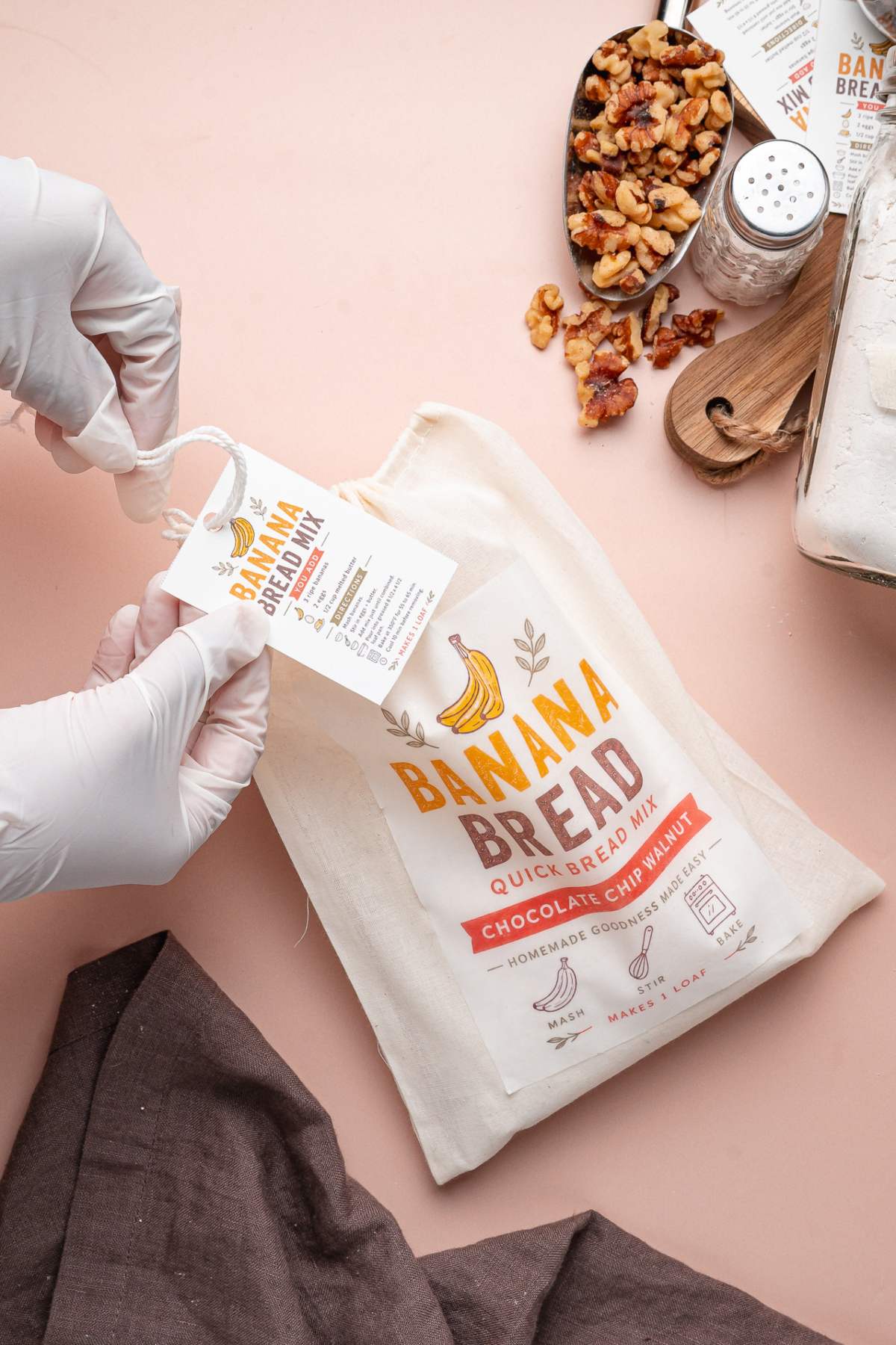 Gloved hands attach a tag to a homemade banana bread mix bag. Walnuts and ingredients are nearby on a pink surface.