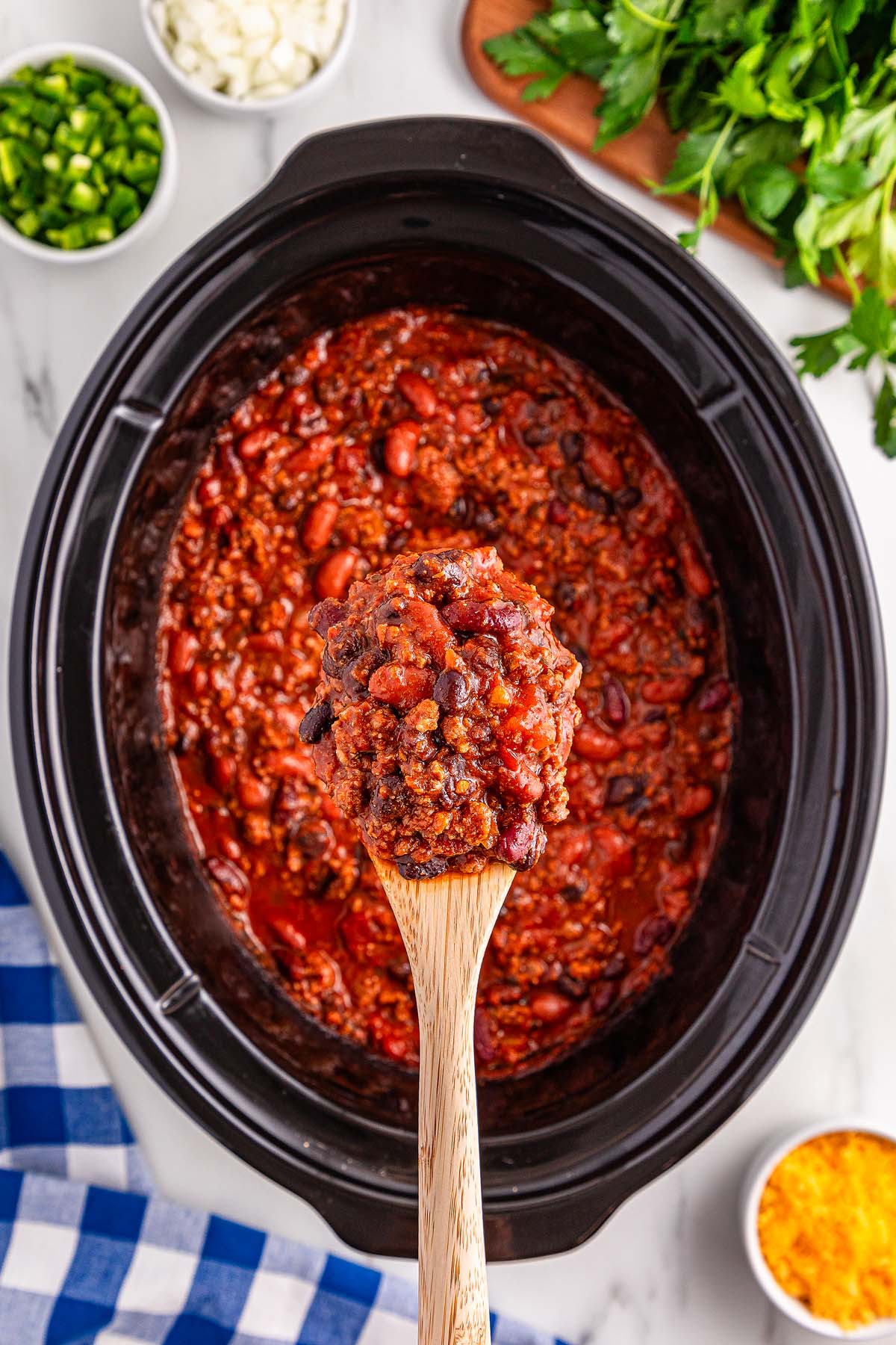 A wooden spoon holding slow cooker chili hovers over a simmering pot, surrounded by bowls of chopped vegetables and herbs.