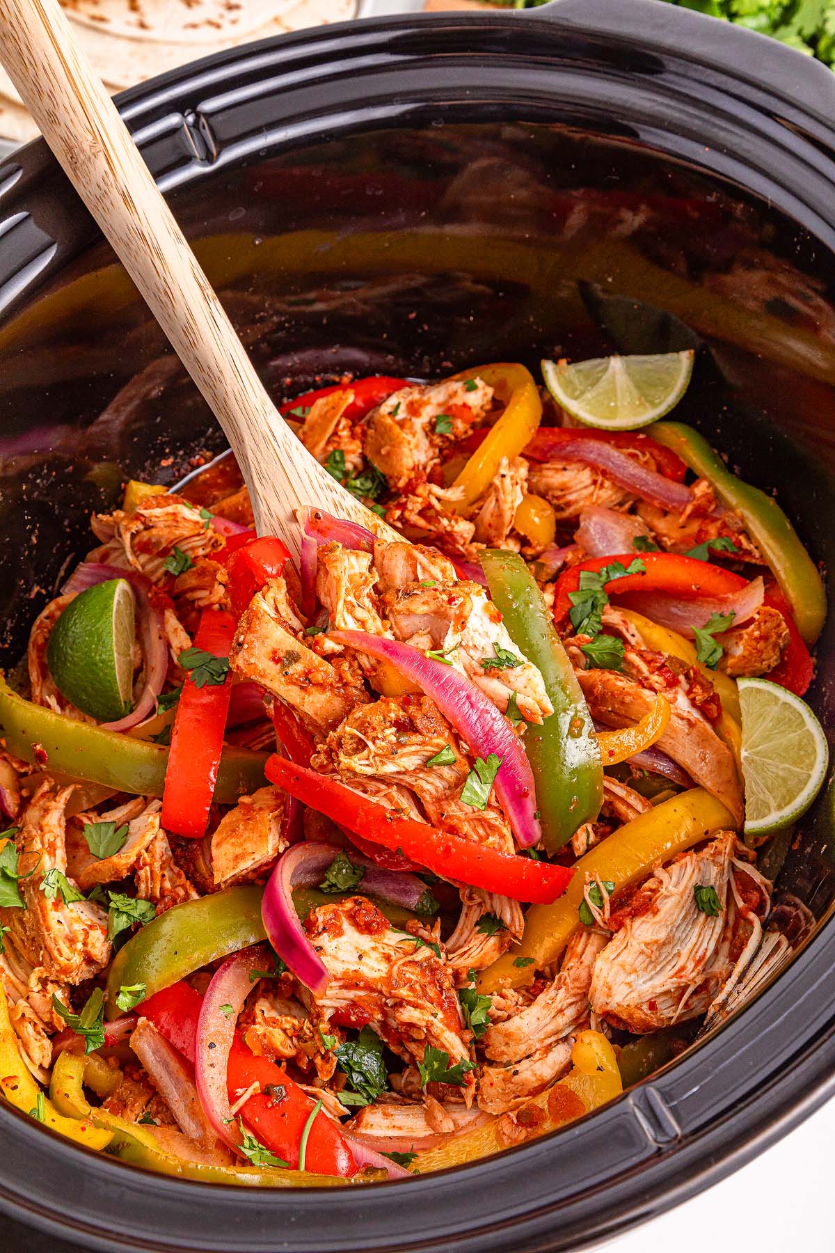 A slow cooker brimming with colorful Slow Cooker Chicken Fajitas, featuring tender chicken, vibrant bell peppers, onions, and fresh lime wedges, all ready to be served with a wooden spoon.