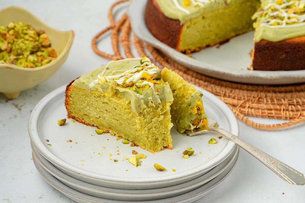 A slice of pistachio ricotta cake with green frosting sits on a plate, topped with chopped pistachios and a delicate white drizzle.