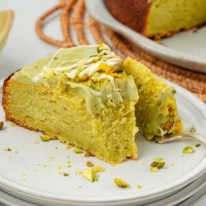 A slice of pistachio ricotta cake with green frosting and chopped pistachios sits on a white plate, accompanied by a fork.