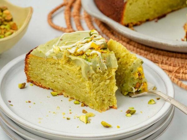 A slice of pistachio ricotta cake with green frosting and chopped pistachios sits on a white plate, accompanied by a fork.