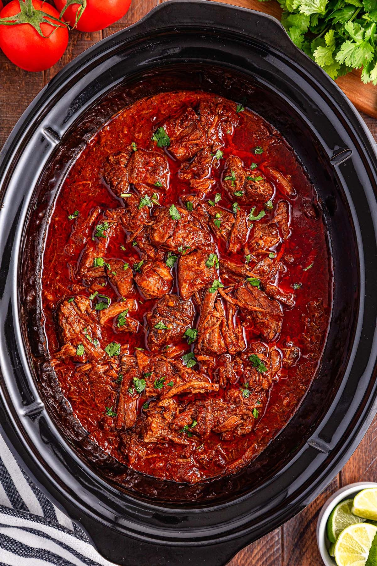 Slow-cooked beef birria in a black crockpot, garnished with chopped cilantro, with tomatoes and limes nearby—a delicious slow cooker birria perfect for savoring rich flavors and tender meat.