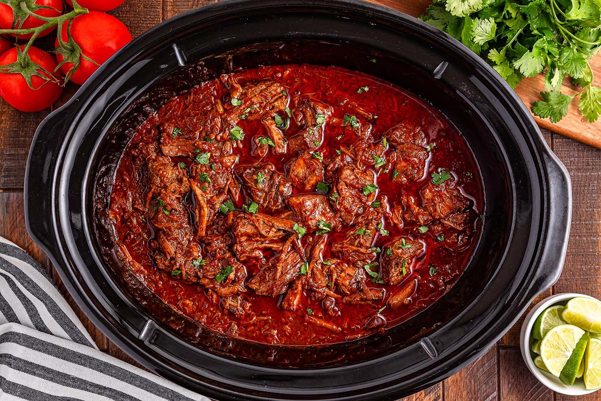 Slow cooker birria filled with shredded beef in rich red sauce, topped with fresh herbs, surrounded by tomatoes and limes.