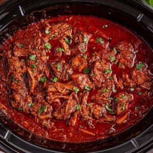 Slow cooker birria made with tender shredded beef, garnished with fresh cilantro, served alongside limes and ripe tomatoes.