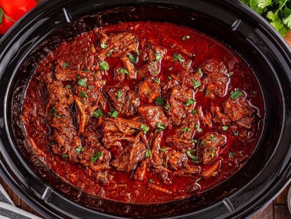 Slow cooker birria made with tender shredded beef, garnished with fresh cilantro, served alongside limes and ripe tomatoes.