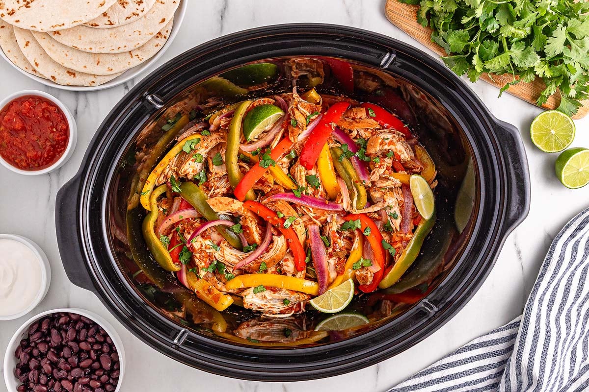 Slow Cooker Chicken Fajitas simmer alongside tender onions and peppers in the slow cooker, surrounded by warm tortillas, hearty beans, zesty salsa, and fresh limes.