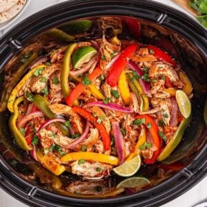 Slow Cooker Chicken Fajitas simmer in a colorful medley of peppers and onions, garnished with fresh lime wedges and cilantro, and served alongside beans, salsa, and warm tortillas.