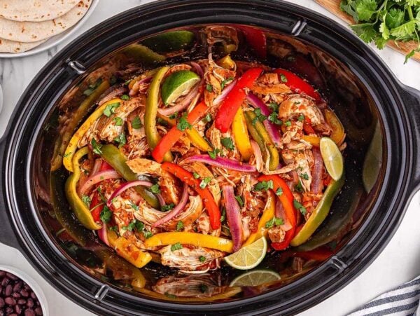 Slow Cooker Chicken Fajitas simmer in a colorful medley of peppers and onions, garnished with fresh lime wedges and cilantro, and served alongside beans, salsa, and warm tortillas.