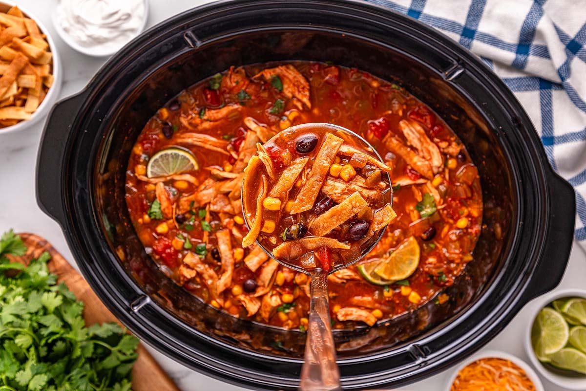 Ladle of Slow Cooker Chicken Tortilla Soup with beans, corn, and lime, in a black slow cooker, with toppings nearby.