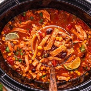 A ladle scoops hearty Slow Cooker Chicken Tortilla Soup, brimming with tender chicken, corn, beans, and fresh lime from a black slow cooker.