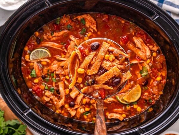 A ladle scoops hearty Slow Cooker Chicken Tortilla Soup, brimming with tender chicken, corn, beans, and fresh lime from a black slow cooker.