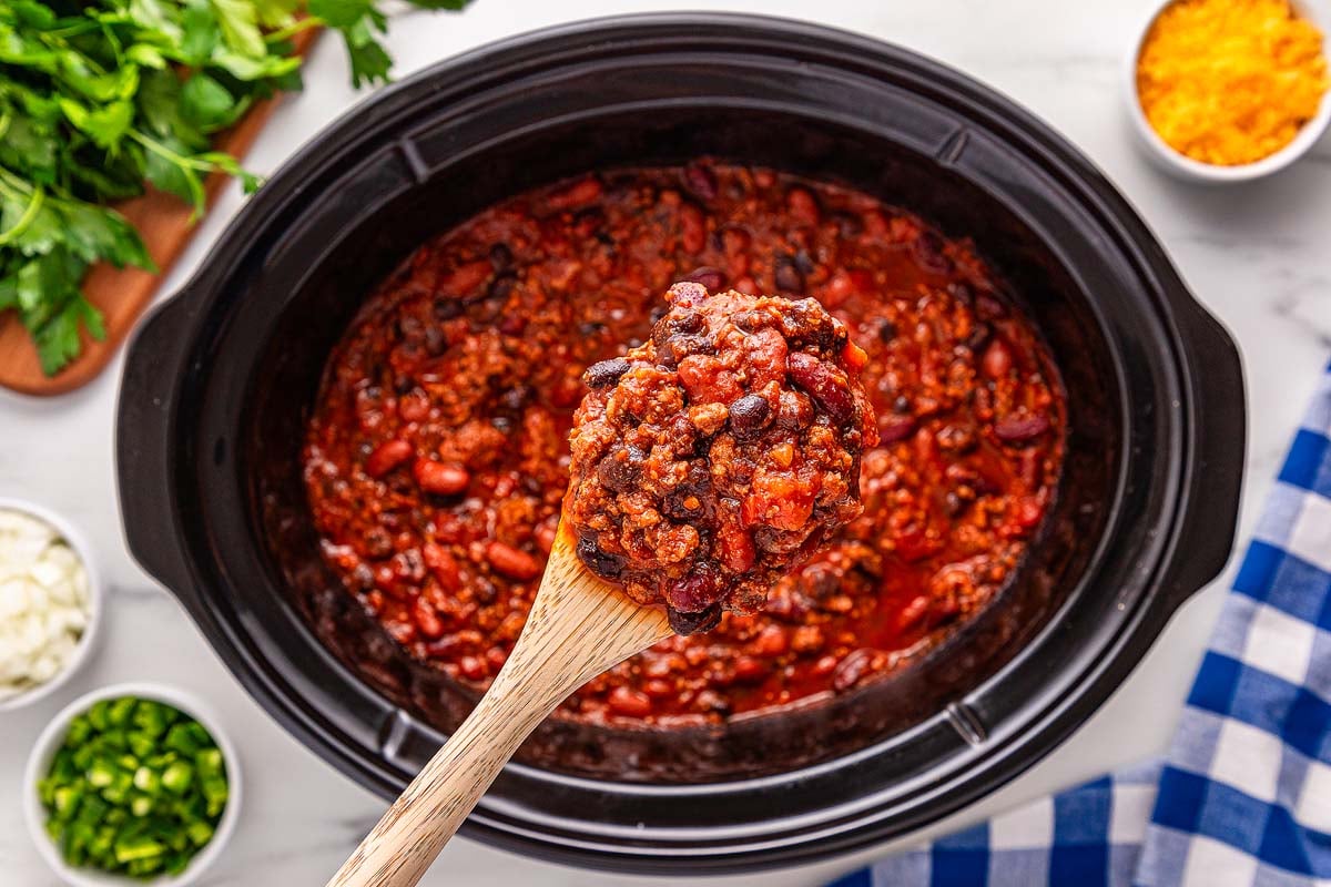 A wooden spoon lifts hearty slow cooker chili with beans from the pot, surrounded by chopped veggies and shredded cheese.