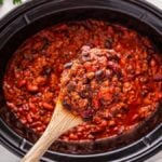 A wooden spoon holds a scoop of slow cooker chili above a slow cooker, with beans and meat simmered in a rich tomato sauce.